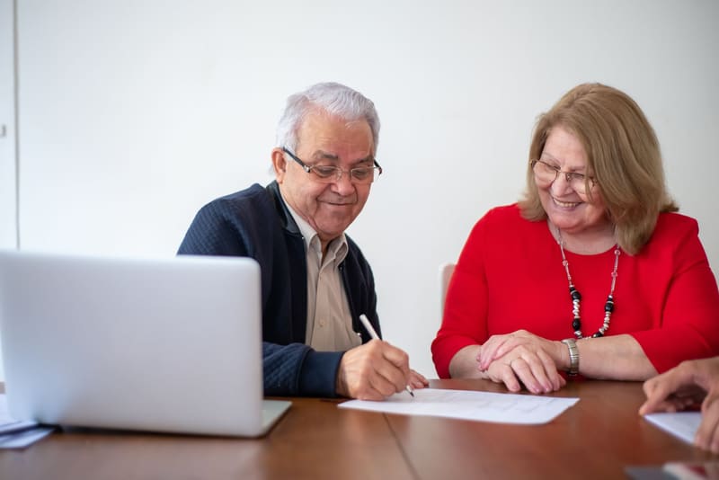 An older man signing papers at a table beside an older woman